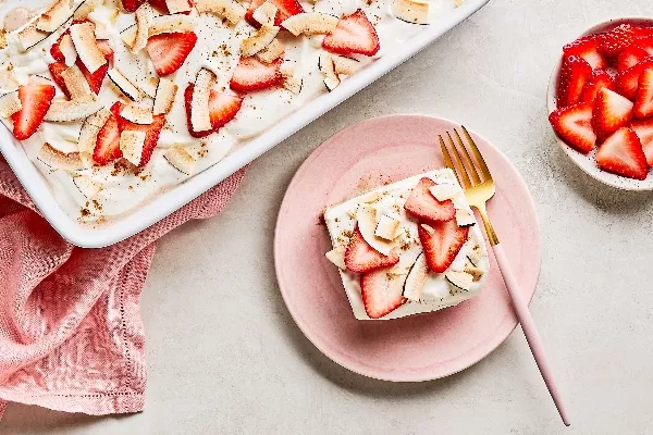 Une part de gâteau tres leches aux fraises et à la noix de coco sur une assiette rose avec une fourchette dorée, déposée à côté d’un moule blanc contenant le même gâteau et d’un petit bol de fraises tranchées, le tout sur un comptoir gris avec une serviette en tissu rose.