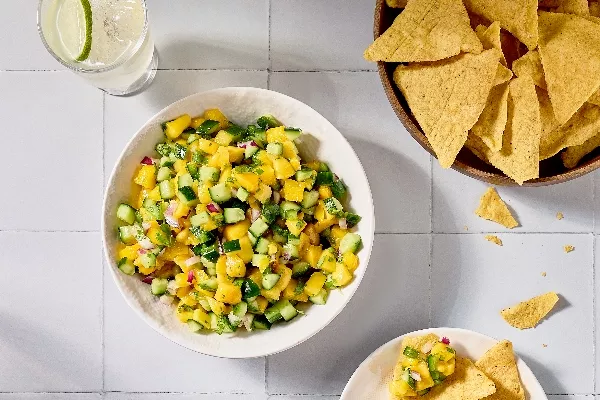 Une image en plongée d'un bol de salsa à la mangue et au concombre sur une surface en carreaux blancs. La salsa comprend également de l' oignon rouge et de la coriandre. Un bol de chips tortilla est placé à côté, avec quelques chips éparpillés autour. On voit également un verre d'eau infusée à la lime.