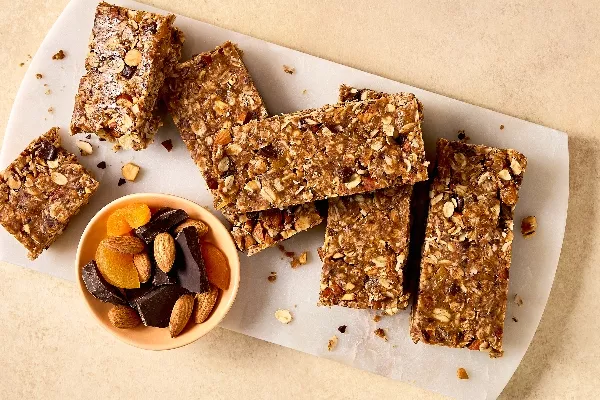 Gros plan sur des barres granola sans cuisson maison contenant de l’avoine, des noix et des fruits secs, disposées sur une planche à découper blanche. Un petit bol avec des amandes, des abricots secs et des morceaux de chocolat noir se trouve à proximité. Les barres sont rectangulaires.