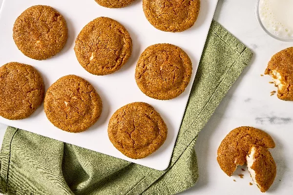 Vue aérienne d’un plateau blanc de biscuits aux épices pour tarte à la citrouille farcis au fromage à la crème, le tout déposé sur un tissu vert. Les biscuits ont une teinte dorée et leur dessus est légèrement craquelé, laissant entrevoir la garniture crémeuse à l’intérieur. L’un des biscuits est ouvert, montrant la garniture au fromage à la crème.