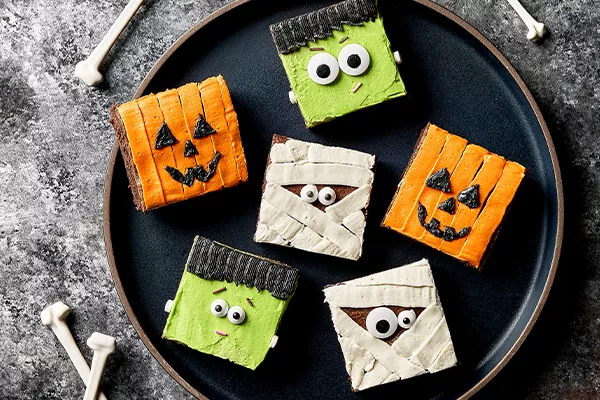 Six brownies carrés décorés pour Halloween comme des citrouilles, des momies et Frankenstein, présentés sur une assiette noire avec des os éparpillés autour.