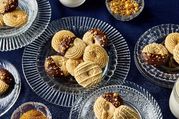 Assortiment de biscuits au beurre décorés et présentés sur des assiettes en verre, mettant en vedette trois styles de décoration : moitié trempé dans du chocolat avec des paillettes dorées, nappé de chocolat blanc et saupoudré de sucre doré. L'arrière-plan est un napperon bleu foncé, avec des paillettes dorées supplémentaires dans un petit bol.