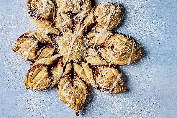 Une pâtisserie Flocon de Neige à la Cannelle Glacé, magnifiquement confectionnée, en forme de flocon avec des couches de pâte torsadées, remplie de cannelle et de sucre. La pâtisserie est nappée d'un léger glaçage et saupoudrée de sucre glace, présentée sur un fond bleu pâle.