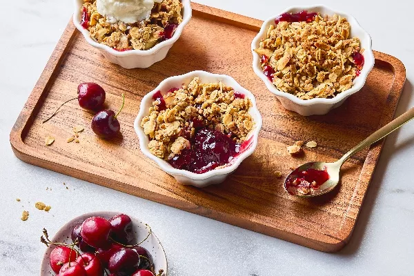 Des portions individuelles de croustade aux cerises et au sarrasin avec un croustillant à l’avoine doré dans des ramequins blancs. Une des croustades est garnie d’une boule de crème glacée à la vanille. Des cerises fraîches sont dispersées sur un plateau de service en bois, un bol de cerises est placé tout près et une cuillère dorée est remplie de garniture juteuse, montrant ainsi la texture et la couleur vibrante du dessert.