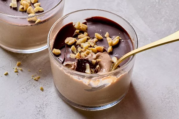 Un plan rapproché d’un pouding crémeux au chocolat et au beurre d’arachides dans un pot de verre, couvert d’une couche de chocolat craquante et d’arachides hachées. Une cuillère dorée est déposée dans le pot, révélant la texture soyeuse et décadente du pouding sous la couche de chocolat. Des arachides émiettées sont étalées sur une surface gris pâle.