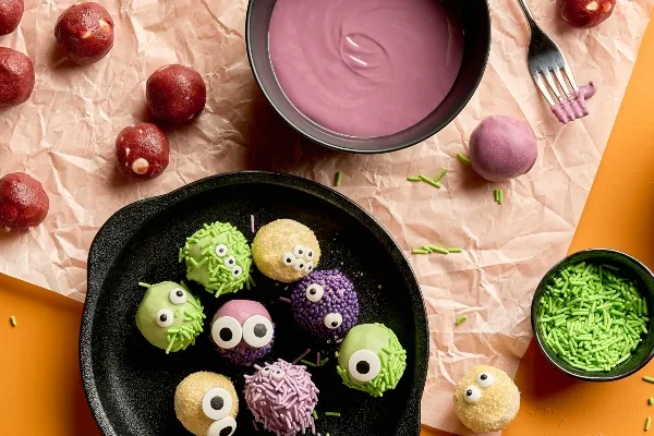 Truffes de pâte à biscuits red velvet sur le thème de l'Halloween, décorées comme des visages de monstres, enrobées de fondant coloré (vert, mauve ou rose), puis roulées dans des décorations en sucre (ou vermicelles de couleur) et garnies d'yeux en bonbons. Un plat noir contient les truffes finies, et des boules de velours rouge non enrobées, un bol de fondant mauve, des vermicelles verts et une fourchette reposent sur du papier parchemin chiffonné à proximité, sur une surface orange vif.