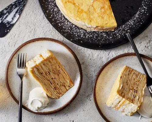 Deux tranches de gâteau crêpe au bourbon sur assiette, servies avec de la crème fouettée et un verre de bourbon.