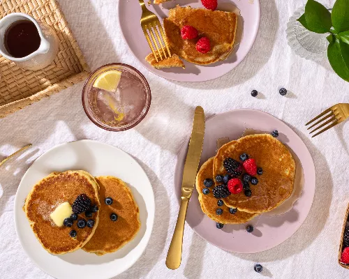 Plan en plongée de trois assiettes de pancakes garnis de petits fruits, disposées sur une table avec des ustensiles dorés, des p