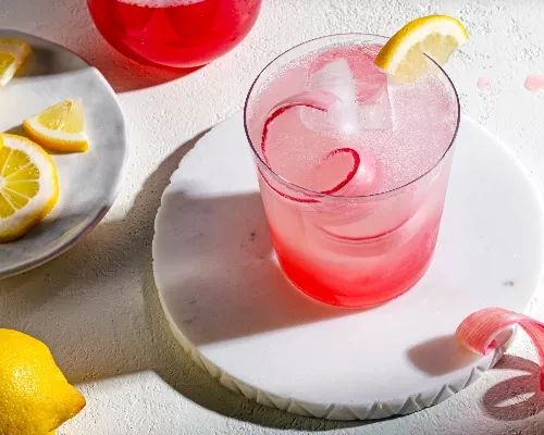 Verre de limonade rose à la rhubarbe sur un dessous de verre garni de citron et de rhubarbe, et une assiette de roues à citron.