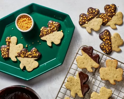 Des biscuits à la menthe en forme de trèfle trempés dans le chocolat et parés de paillettes dorées, disposés sur un plateau vert