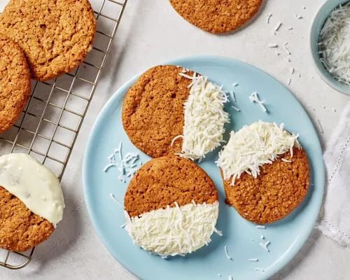 Des biscuits au gingembre à moitié trempés dans le chocolat blanc et de la noix de coco sur une assiette bleue et sur une grille