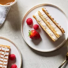  Deux morceaux de gâteau Mille-Feuille sur des assiettes, présentés avec des baies, une tasse de café et une fourchette dorée.