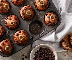 Muffins végétaliens à la banane et aux pépites de chocolat dans un moule à muffins, avec un muffin retiré et ouvert en deux, et un bol de pépites de chocolat.