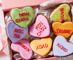 Des biscuits en sucre en forme de cœur décorés de glaçage royal avec des messages romantiques écrits dans le glaçage, présentés sur des carreaux blancs avec des bols de glaçage royal et une poche à douille.
