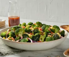 Un saladier de salade de brocoli avec une vinaigrette au gochujang est présenté sur un comptoir de cuisine, avec une carafe de vinaigrette, des assiettes et des cuillères de service en bois.
