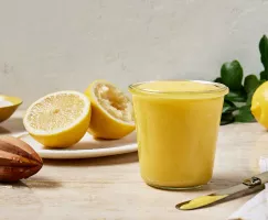 Un pot en verre rempli de lemon curd est présenté avec un citron coupé en deux, dont une moitié est pressée, un presse-citron et un bol de sucre granulé.