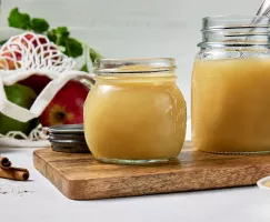 Deux bocaux en verre de compote de pommes maison sur une planche à découper en bois sur une surface blanche, un sac en maille de pommes, des bâtons de cannelle et un bol de Cassonade Dorée.