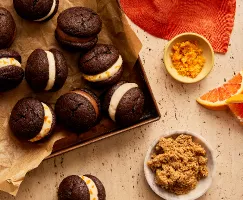 Des mini whoopie pies au chocolat avec diverses garnitures sur une plaque de cuisson doublée de papier parchemin, présentés avec un bol de sucre brun foncé, un bol de zeste d'orange et des quartiers d'orange.