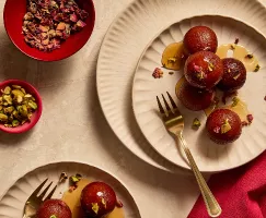  Des gulab jamuns arrosés de sirop et garnis de morceaux de pistaches et de pétales de rose, répartis entre deux assiettes et accompagnés de fourchettes en or.