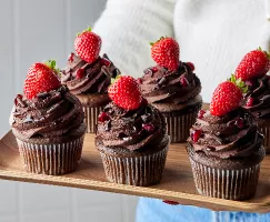 Six cupcakes au chocolat et au vin rouge garnis de ganache, de copeaux de chocolat, de grenade et de fraises sur un plateau en bois tenu par une femme en pull blanc et en jean