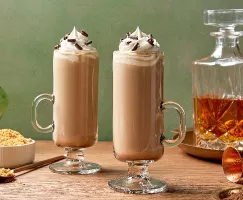 Deux grands verres de latte au whisky irlandais surmontés de crème fouettée et de copeaux de chocolat, présentés sur une table en bois avec un accompagnement de sucre brun, une carafe de whisky ambre en arrière-plan.