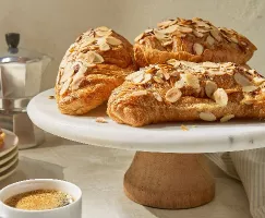 Quatre croissants aux amandes cuits à la friteuse à air, garnis d'amandes tranchées sur un présentoir à gâteaux blanc, avec une tasse d'espresso et une cafetière à l'arrière-plan.