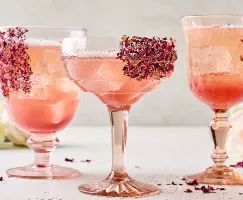 Trois verres à tige uniques de spritz litchi et rose sur glaçons. Des pétales de rose séchés garnissent les verres qui sont présentés dans un cadre neutre avec des roses fraîches en arrière-plan et des pétales de rose séchés dispersés autour.