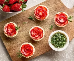 Cinq mini-tartelettes fraises et romarin sur une planche à découper en bois, présentées avec un bol de brins de romarin, un bol de fraises fraîches et une sixième mini-tartelette déposée sur le comptoir.