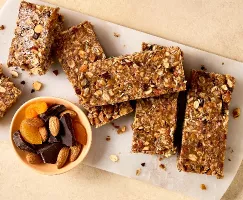 Gros plan sur des barres granola sans cuisson maison contenant de l’avoine, des noix et des fruits secs, disposées sur une planche à découper blanche. Un petit bol avec des amandes, des abricots secs et des morceaux de chocolat noir se trouve à proximité. Les barres sont rectangulaires.