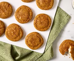 Vue aérienne d’un plateau blanc de biscuits aux épices pour tarte à la citrouille farcis au fromage à la crème, le tout déposé sur un tissu vert. Les biscuits ont une teinte dorée et leur dessus est légèrement craquelé, laissant entrevoir la garniture crémeuse à l’intérieur. L’un des biscuits est ouvert, montrant la garniture au fromage à la crème.