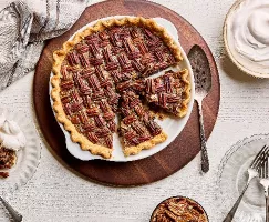 Une tarte au pacane dans un plat à gratin, montrant trois tranches coupées, dont une servie sur une assiette avec de la crème fouettée, sur une table en bois blanc avec un bol de crème fouettée et un bol de pacanes.