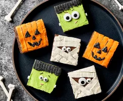 Six brownies carrés décorés pour Halloween comme des citrouilles, des momies et Frankenstein, présentés sur une assiette noire avec des os éparpillés autour.