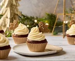 Des petits gâteaux au pain d'épices festifs surmontés d'un tourbillon de glaçage blanc crémeux et d'un mini biscuit au pain d'épices. Les petits gâteaux sont disposés sur une surface de bois avec des décorations de Noël en arrière-plan, dont un sapin de Noël doré et des lumières féériques scintillantes.