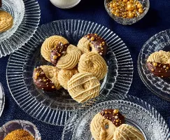 Assortiment de biscuits au beurre décorés et présentés sur des assiettes en verre, mettant en vedette trois styles de décoration : moitié trempé dans du chocolat avec des paillettes dorées, nappé de chocolat blanc et saupoudré de sucre doré. L'arrière-plan est un napperon bleu foncé, avec des paillettes dorées supplémentaires dans un petit bol.
