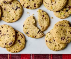Des biscuits à la canneberge et à la pistache disposés sur une surface de marbre blanc. Les biscuits tranchés et cuits au four présentent des morceaux visibles de canneberges séchées et de pistaches, ajoutant texture et couleur. Un biscuit est coupé en deux, révélant une mie tendre. Un tissu à carreaux rouges ajoute une touche festive au bas de l'image.