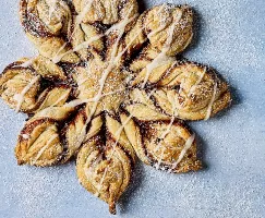 Une pâtisserie Flocon de Neige à la Cannelle Glacé, magnifiquement confectionnée, en forme de flocon avec des couches de pâte torsadées, remplie de cannelle et de sucre. La pâtisserie est nappée d'un léger glaçage et saupoudrée de sucre glace, présentée sur un fond bleu pâle.