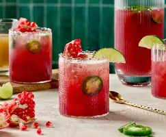 Trois verres de Fizz Épicé à la Grenade et au Gingembre, d'une couleur rouge vibrante avec des tranches de jalapeño frais. Les boissons sont garnies de quartiers de lime et de graines de grenade. Une carafe remplie de la même boisson se trouve en arrière-plan, aux côtés de quartiers de lime, de morceaux de grenade et d'une petite carafe de sirop de gingembre. La scène se déroule sur un fond de tuiles vert émeraude.
