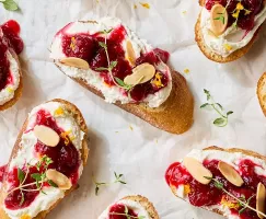 Un gros plan sur plusieurs crostinis garnis de feta fouettée, de chutney de canneberges, d'amandes effilées et de thym frais.