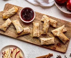 Une planche à découper en bois est recouverte d'un tas de rouleaux impériaux dorés et croustillants. Un petit bol de confiture de fraises se trouve à proximité. Des tranches de pommes et des bâtons de cannelle sont éparpillés autour de la planche, suggérant que les rouleaux impériaux sont farcis aux pommes et à la cannelle.