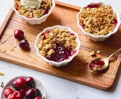 Portions individuelles de croustillant cerise et sarrasin dans des ramequins blancs, garnies d'une croustillante avoine dorée, dont une avec une boule de crème glacée à la vanille. Des cerises fraîches sont dispersées sur le plateau de service en bois, avec un bol de cerises à proximité et une cuillère dorée contenant une partie de la garniture juteuse, mettant en évidence la couleur et la texture vibrantes du dessert.