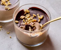 Gros plan d'un crémeux pouding au chocolat et beurre de cacahuète dans un pot de verre, garni d'une couche de chocolat lustrée et de cacahuètes hachées. Une cuillère dorée repose à l'intérieur, révélant la texture lisse et gourmande sous la coque de chocolat. Des cacahuètes émiettées sont éparpillées sur la surface gris clair.