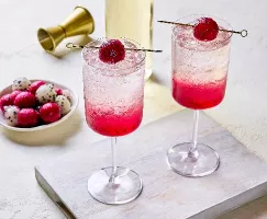 Deux verres de spritzer vibrant au fruit du dragon servis sur glace, avec un dégradé rose provenant du fruit au fond. Chaque verre est garni d'une boule de fruit du dragon en brochette. Une assiette de boules de fruit du dragon rouges et blanches, un jigger doré et une bouteille sont visibles à l'arrière-plan.