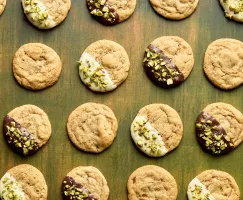Biscuits sans pépites de chocolat disposés en quadrillage sur une surface en bois vert. Certains biscuits ne sont pas décorés, tandis que d’autres sont à moitié trempés dans du chocolat blanc ou noir et saupoudrés de pistaches hachées. 