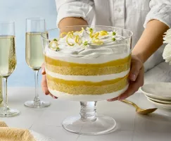 Deux mains qui tiennent un bol de service en verre rempli d’une bagatelle au citron et à la fleur de sureau avec ses couches de gâteau éponge, de curd au citron et de crème fouettée. La bagatelle est couronnée de rubans de zeste de citron et de délicates fleurs blanches. Des flûtes à champagne, une bouteille et des roses blanches dans un vase en verre sont visibles en arrière-plan.