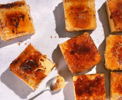 Une prise de vue de dessus de plusieurs petites tranches carrées de gâteau au fromage caramélisé avec une garniture de sucre brûlé, éparpillées sur une surface blanche avec une cuillère prenant une bouchée d'un morceau.
