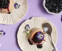 Vue de dessus de deux portions de panna cotta à l'Earl Grey garnies de compote de bleuets et décorées de fleurs comestibles violettes et blanches, servies sur des assiettes crème à bord festonné sur un fond lavande avec des fleurs éparpillées et une cuillère.
