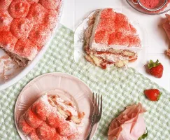 Un plat blanc contenant un tiramisu aux fraises et à la crème repose sur un tissu vichy vert, sur une surface en carreaux blancs. Deux portions coupées sont présentées sur des assiettes roses et en verre clair, avec des fraises fraîches et des roses roses tout autour.