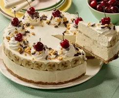 Une tarte glacée façon banana split couronnée de tourbillons de crème fouettée, de cerises, de noix concassées et de copeaux de chocolat déposée sur une assiette blanche. Une part est soulevée pour révéler des couches de rondelles de bananes et de garniture glacée sur une croûte de biscuits Graham. En arrière-plan, on voit une pile d’assiettes pastel avec des fourchettes et un bol de cerises fraîches sur une nappe vert pâle.