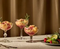 Trois coupes à cocktail remplies d’un dessert glacé composé de couches de slush dorée et rouge rubis, garnies de brins de romarin frais, posées sur une surface claire et texturée devant un rideau brun chaud. À côté, une assiette contient des limes entières, des quartiers de lime, des cerises et du romarin, ainsi qu’un petit pichet en verre rempli de sirop de cerise noire.