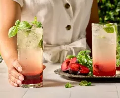 Une personne portant un ensemble en lin de couleur crème tient un grand verre de boisson étagée à la prune garni de feuilles fraîches de basilic et de menthe. À la base du verre, une couche de fruit rouge foncé et, pour le reste, une boisson pétillante légèrement colorée. Sur la table se trouvent d’autres verres, des herbes fraîches et des prunes coupées en deux sur un plateau argenté, le tout sur une toile de fond d’un vert vibrant.